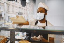 A baker wearing a face mask serves fresh bread to a customer in a modern bakery.