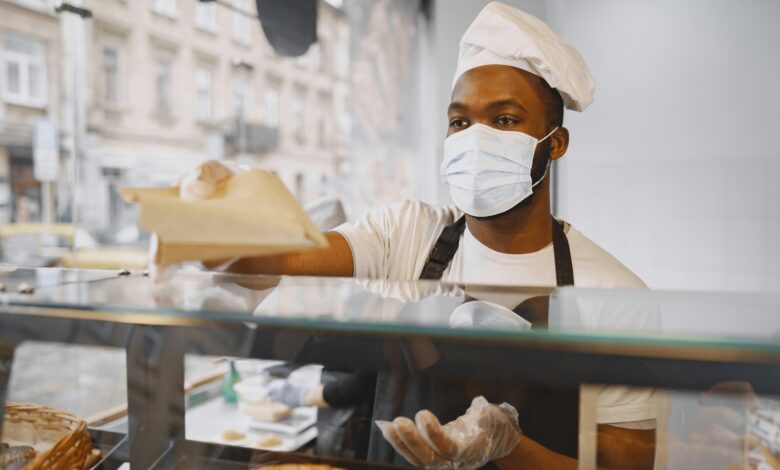 A baker wearing a face mask serves fresh bread to a customer in a modern bakery.