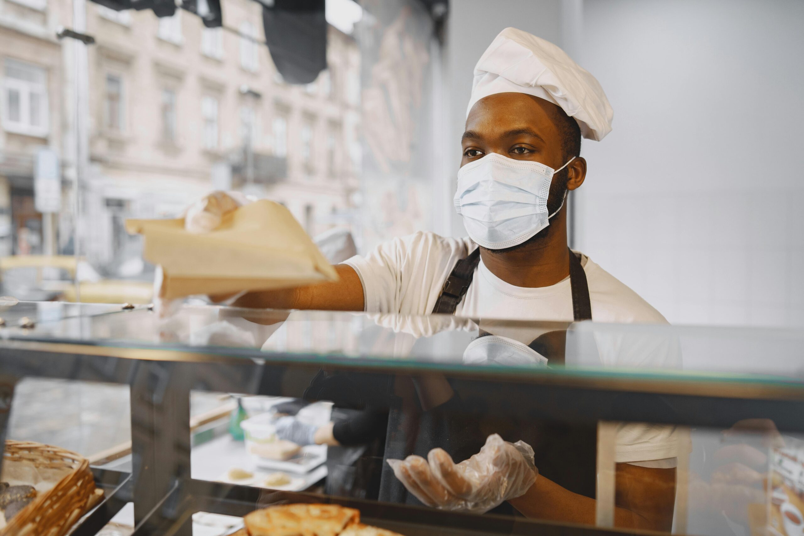 A baker wearing a face mask serves fresh bread to a customer in a modern bakery.